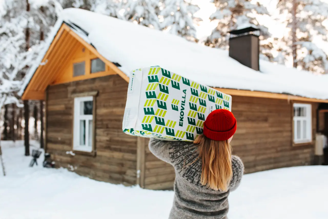Kvinna bär en Ekovilla-säck mot ett snötäckt rött trähus